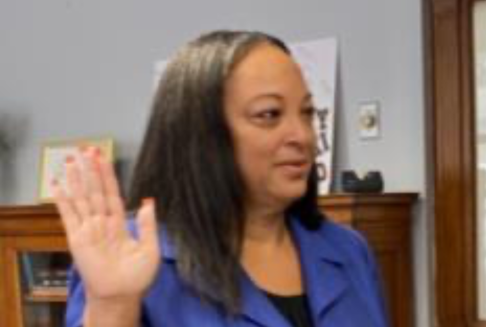 A black woman in a blue jacket with one hand raised to be sworn in.